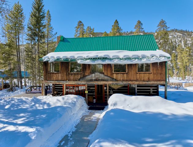 A quaint wooden cabin sits in a snowy landscape, with clear blue skies and sunlit snow-covered trees surrounding it.