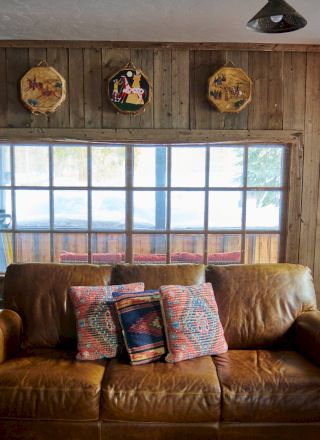 A cozy living room with warm wood tones, a plaid sofa and chairs, a coffee table, soft lighting, and large windows letting in natural light.
