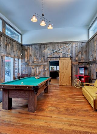 A modern game room with a pool table, lounge seating, warm wooden floors, brick and glass walls, and art-lit decor.