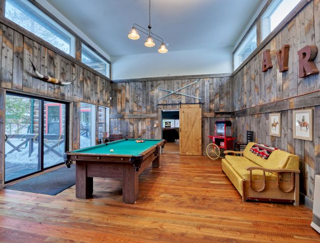 A modern game room with a pool table, lounge seating, warm wooden floors, brick and glass walls, and art-lit decor.