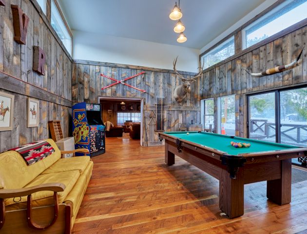 A cozy game room with a pool table, warm lighting, a kitchen, and comfortable seating in the background, perfect for socializing and play.