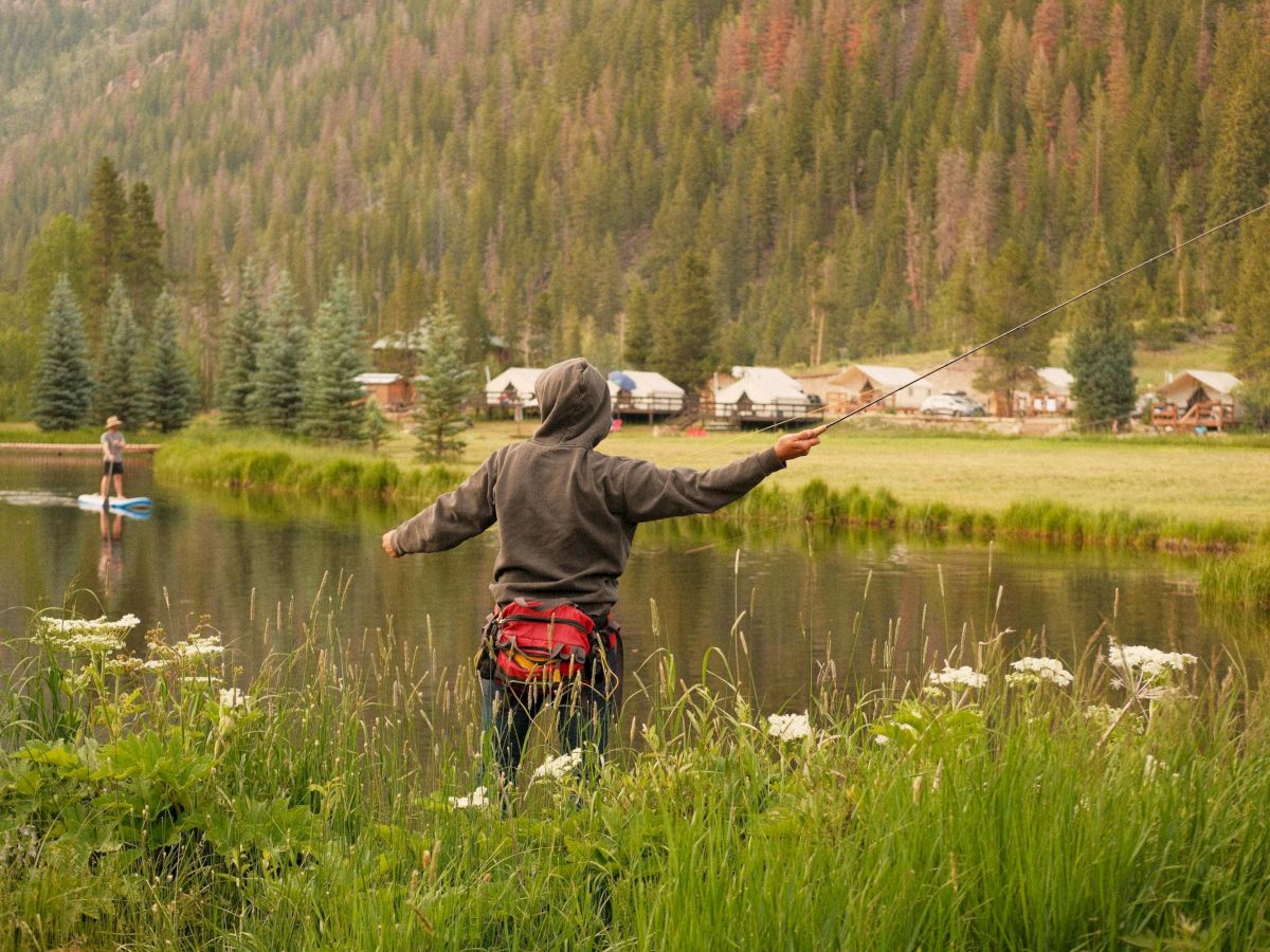 A person in a hoodie stands near a pond with arms outstretched, grassy banks, trees, and a distant row of houses in a forested area.