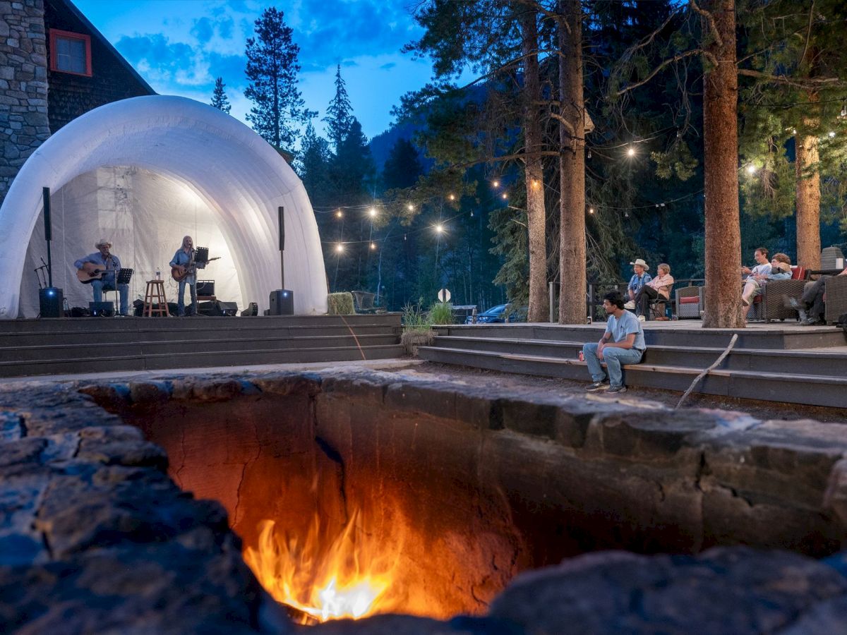 A cozy outdoor concert by a glowing dome stage, with a fire pit in the foreground and people seated among tall trees at dusk.