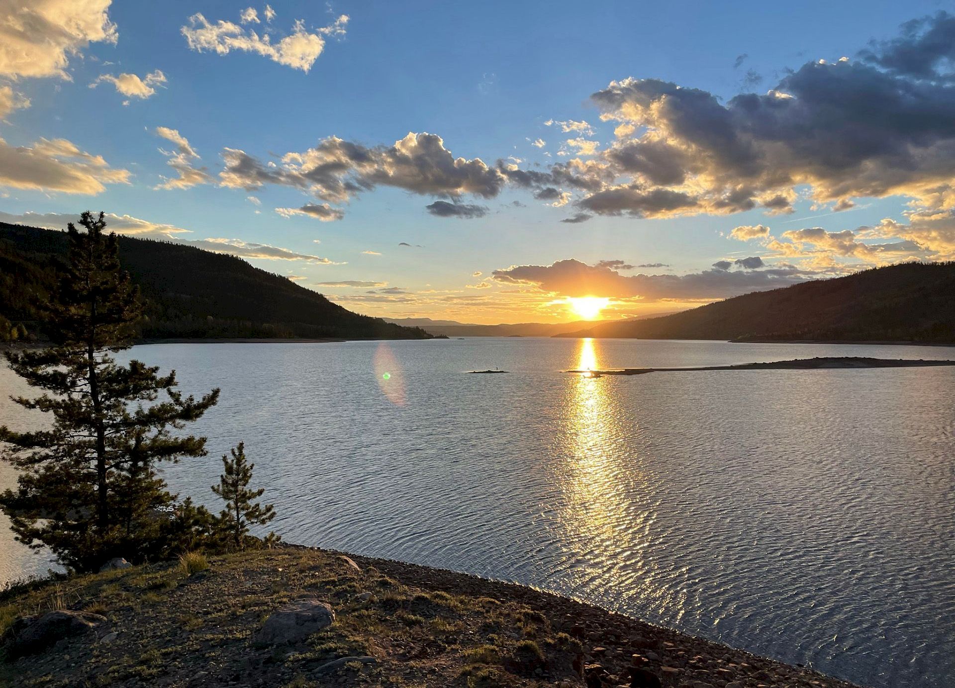 Sunset over a calm lake with rocky shore, a few trees, and golden light reflecting off the water, clouds glowing in the sky.