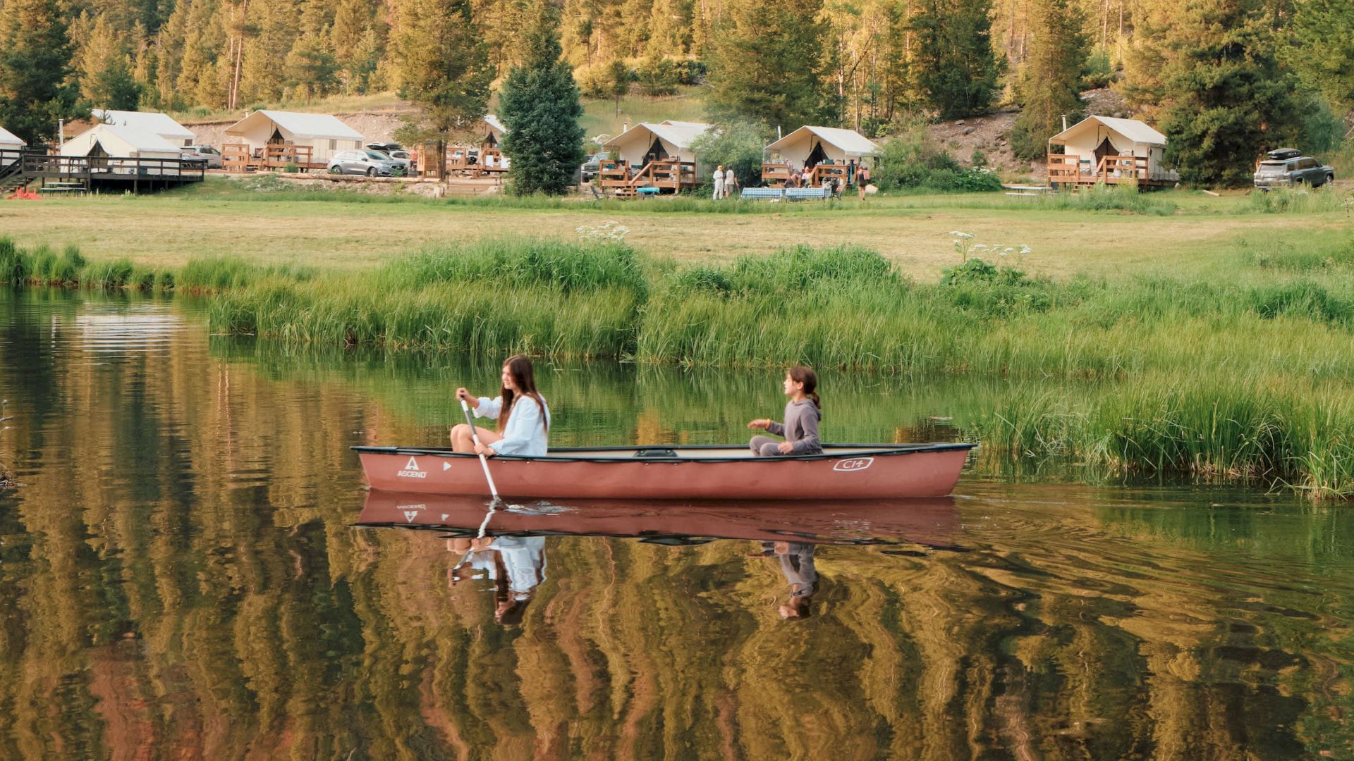 A woman rows a canoe on a calm lake while a child sits onboard, with riverside cabins and a forested hillside behind.