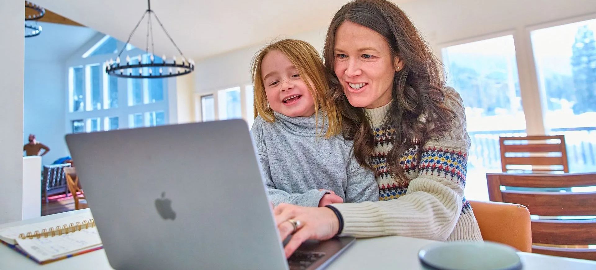 A smiling woman and child sit together at a laptop, looking at the screen in a bright, modern kitchen/dining space.