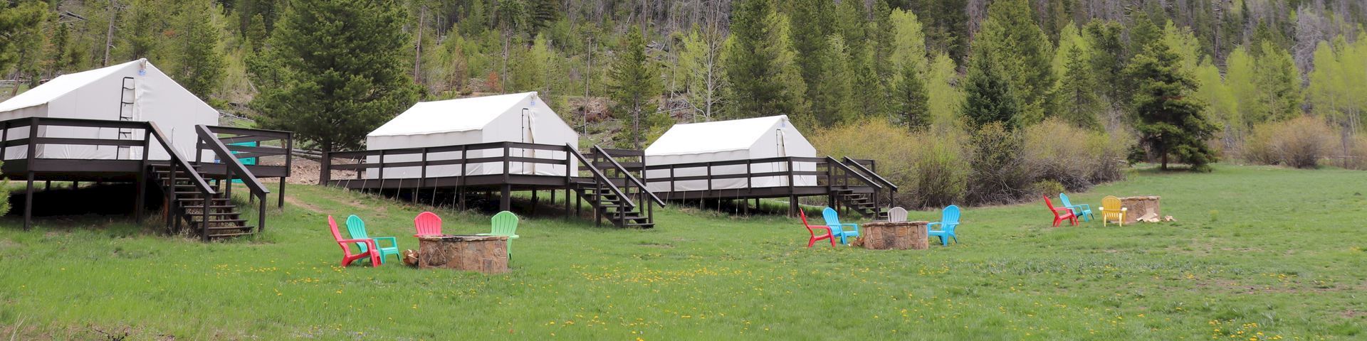 A grassy field with several white yurts on wooden porches, pine trees and forested hills in the background, and colorful chairs scattered about.