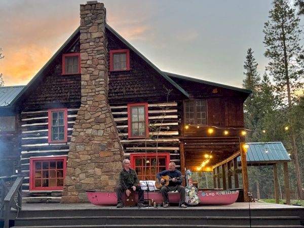 A rustic log cabin with red-framed windows, a tall stone chimney, and two people playing guitar on the porch, string lights glowing gently.