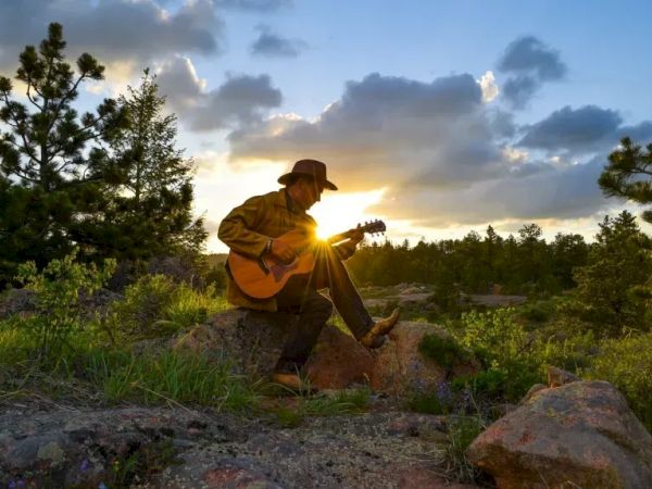 A person wearing a hat sits on a rock, playing an acoustic guitar in a sunlit, forested landscape with clouds and trees. ending with a period.