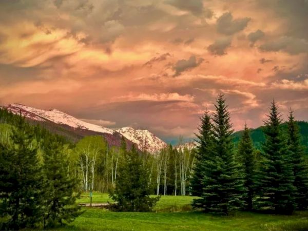 A scenic mountain valley with evergreen trees, snow-dusted peaks, and a dramatic pink-orange sunset sky overhead.