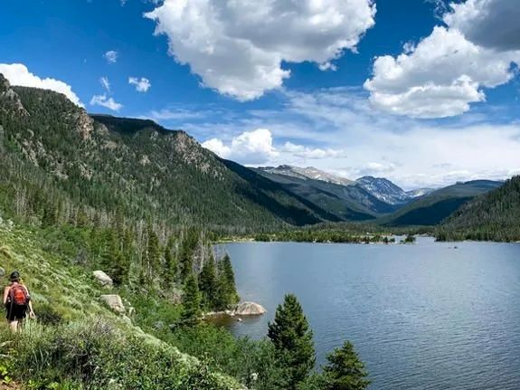 Two hikers trek along a forested lakeside trail, with blue skies, fluffy clouds, and rugged mountains reflected in the calm alpine water.