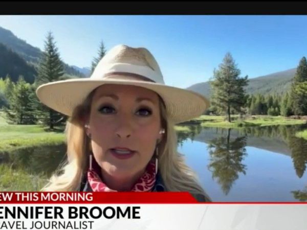 A female travel journalist Jennifer Broome reports live outdoors, with a hat, beside a lake and forested mountains in the background.