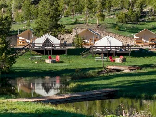 Camp huts by a lake with a wooden deck, tents, trees, and a peaceful grassy area; reflection on the water adds charm, all outdoors.