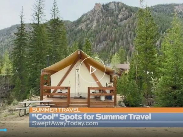 A wooden tent cabin on stilts sits among pine trees with mountains in the background, part of a summer travel show.