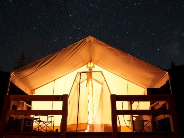 A glowing tent at night, lit from inside, with wooden railings and a starry sky above it.