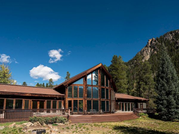 A rustic wooden lodge-style house with large windows, nestled among trees and mountains under a bright blue sky.