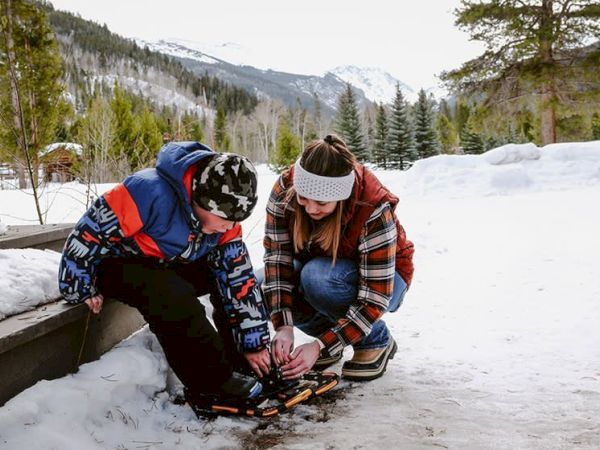 Two kids in winter gear are tying or adjusting skates on a snowy path by a wooden bench, with trees and mountains in the background.