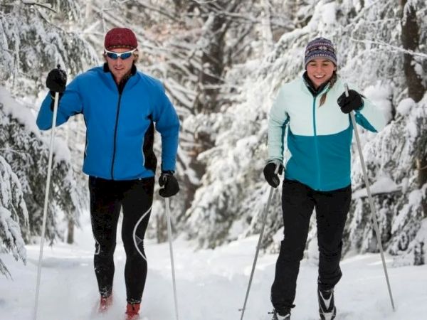 Two people cross-country skiing through a snowy forest, wearing bright jackets and gloves, using poles, in winter gear.