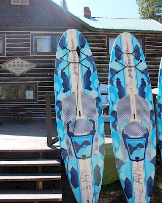 Two blue and white stand-up paddleboards stacked on a wooden deck in front of a log cabin, with a sign and windows visible, under a sunny sky.