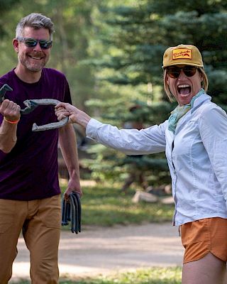Two people outdoors: a man in a purple shirt and tan shorts, and a woman in a light shirt, orange shorts, yellow cap, smiling with snacks, in a forest park.