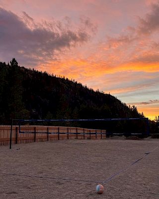 A sunset over a sandy volleyball court, with a fence and distant trees against a colorful orange-pink sky, calm, serene, and empty.