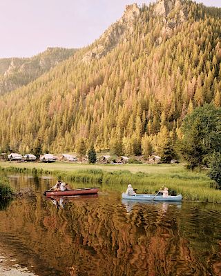 A serene lakeside scene with two small boats drifting on calm water, framed by a forested mountainside and a cluster of lakeside houses, reflective and tranquil.