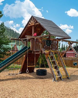 A wooden playground with a slide, climbing ladder, a tire swing, and a shaded platform, set among trees under a bright, partly cloudy sky.