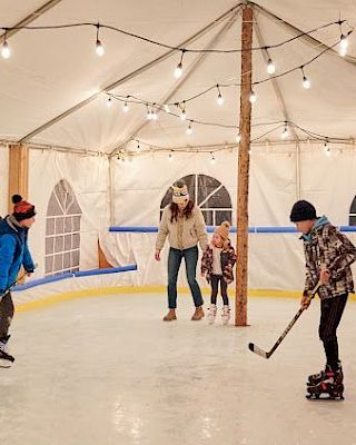 A cozy ice rink inside a tent with string lights; kids and adults skate while a helper watches and a player swings a hockey stick.