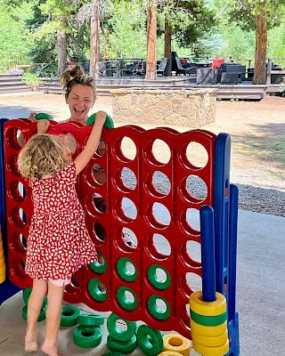Two kids play Connect Four on a blue and red plastic game outdoors, with green circular pieces and tall yellow/blue supports nearby.