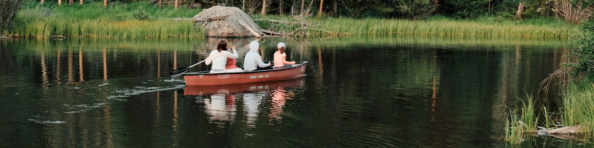 Two people are in a small red boat on a calm lake, surrounded by tall pine trees and a forest, with a rocky shoreline nearby.