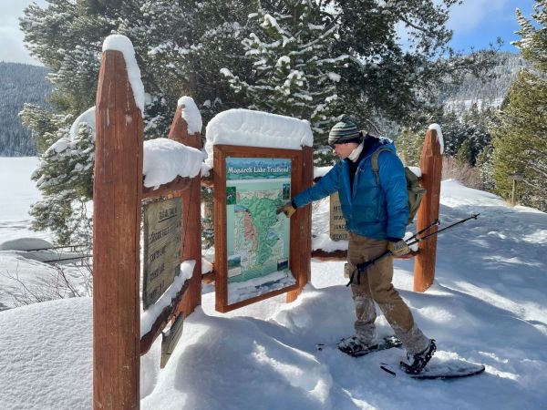 A hiker in a blue vest reads an outdoor trail map at a snowy, forested information sign on a bright winter day.