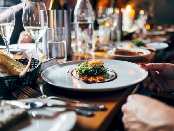 A fine dining scene with a plated dish at center, surrounded by wine glasses, a bread basket, and diners&rsquo; hands reaching for food.