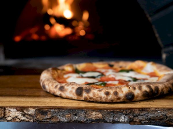 A wood-fired pizza rests on a wooden board in a warm oven glow, with bubbling cheese and toppings ready to slice.