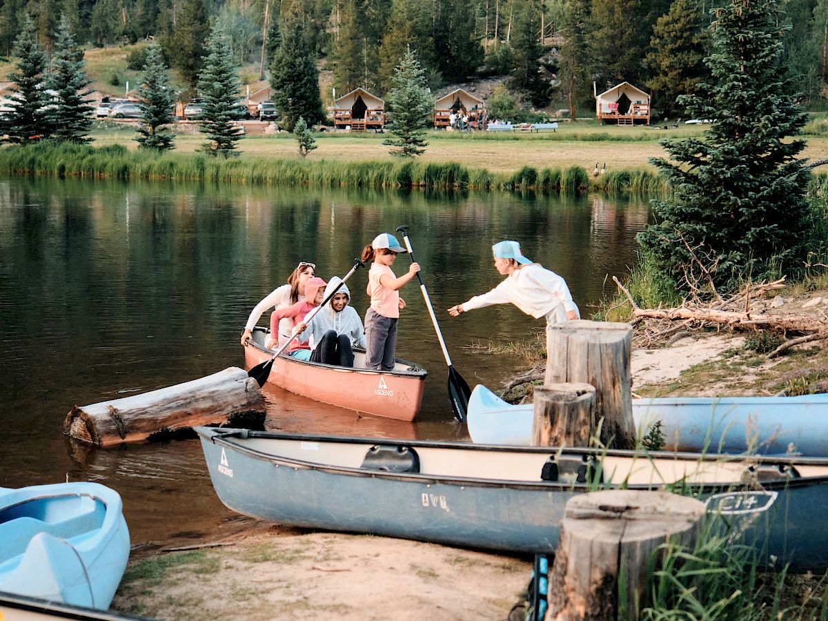 A group of people loading a small orange boat at a lakeside dock, surrounded by pine trees and distant cabins; calm water reflects the scene.