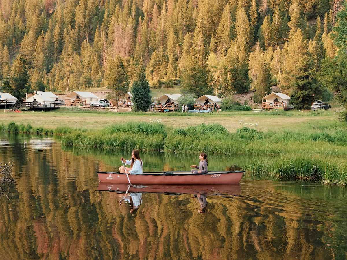 Two people fishing from a red canoe on a calm lake, with a forested mountain and a few cabins in the background, reflected on the water.