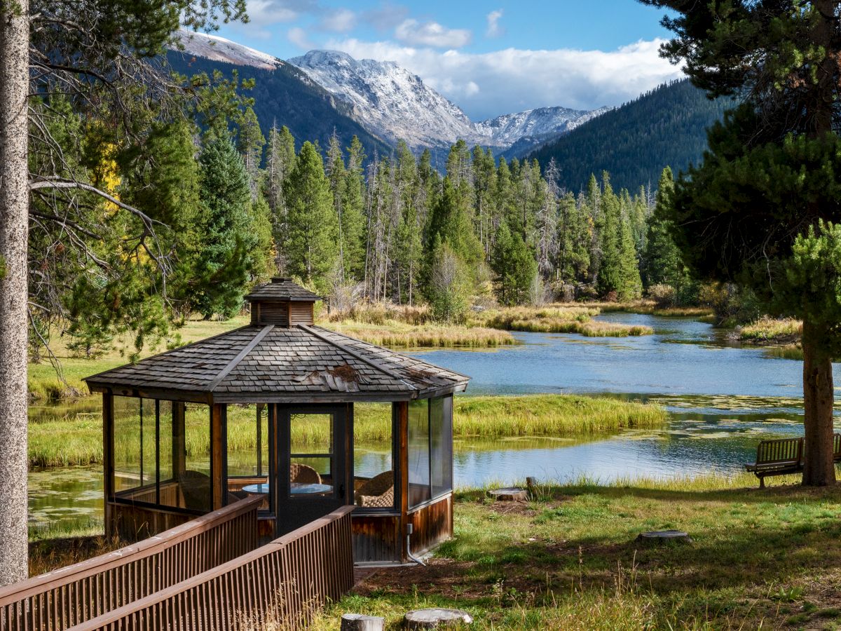 A serene lakeside scene with a wooden gazebo by the water, pine trees, and distant mountains under a blue sky, inviting quiet reflection.