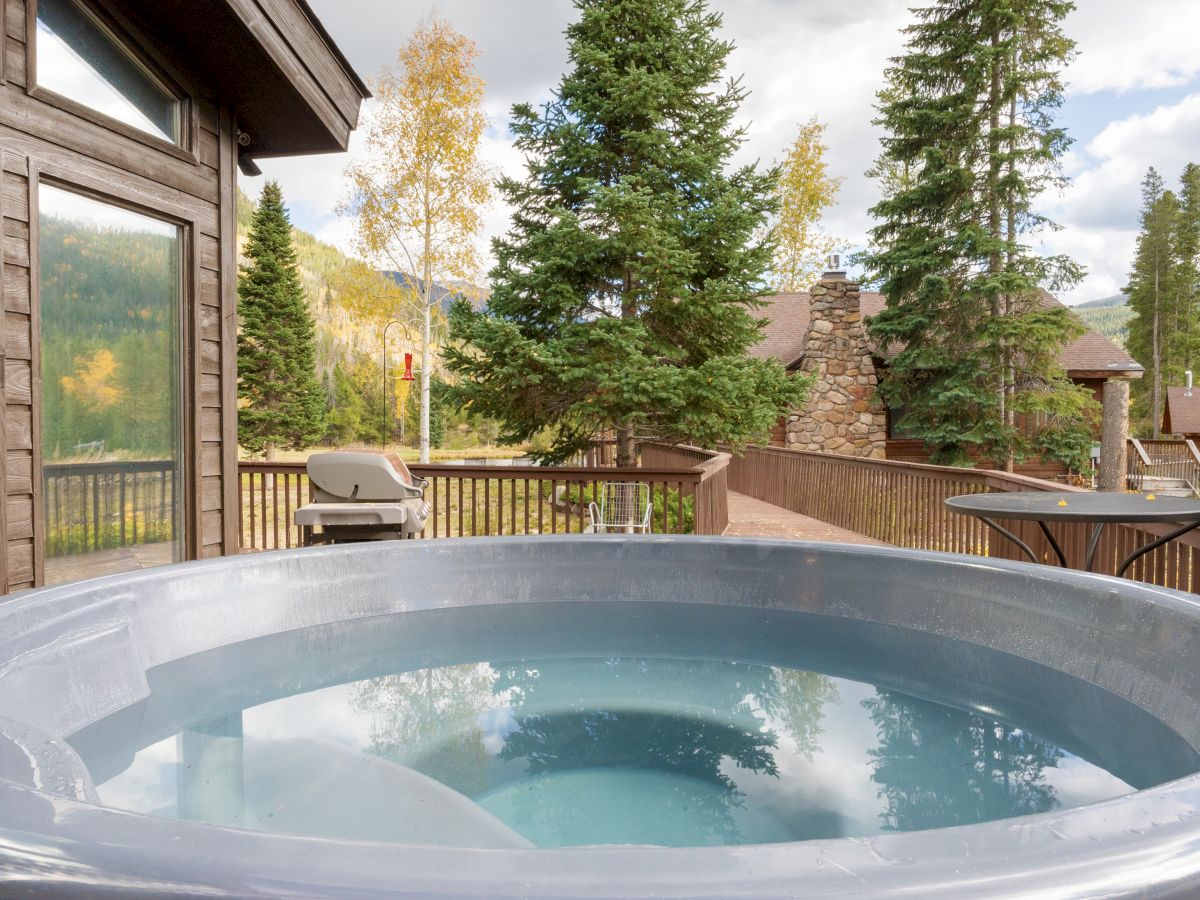 A hot tub sits on a wooden deck outside a cabin, with pine trees and distant mountains visible under a partly cloudy sky.