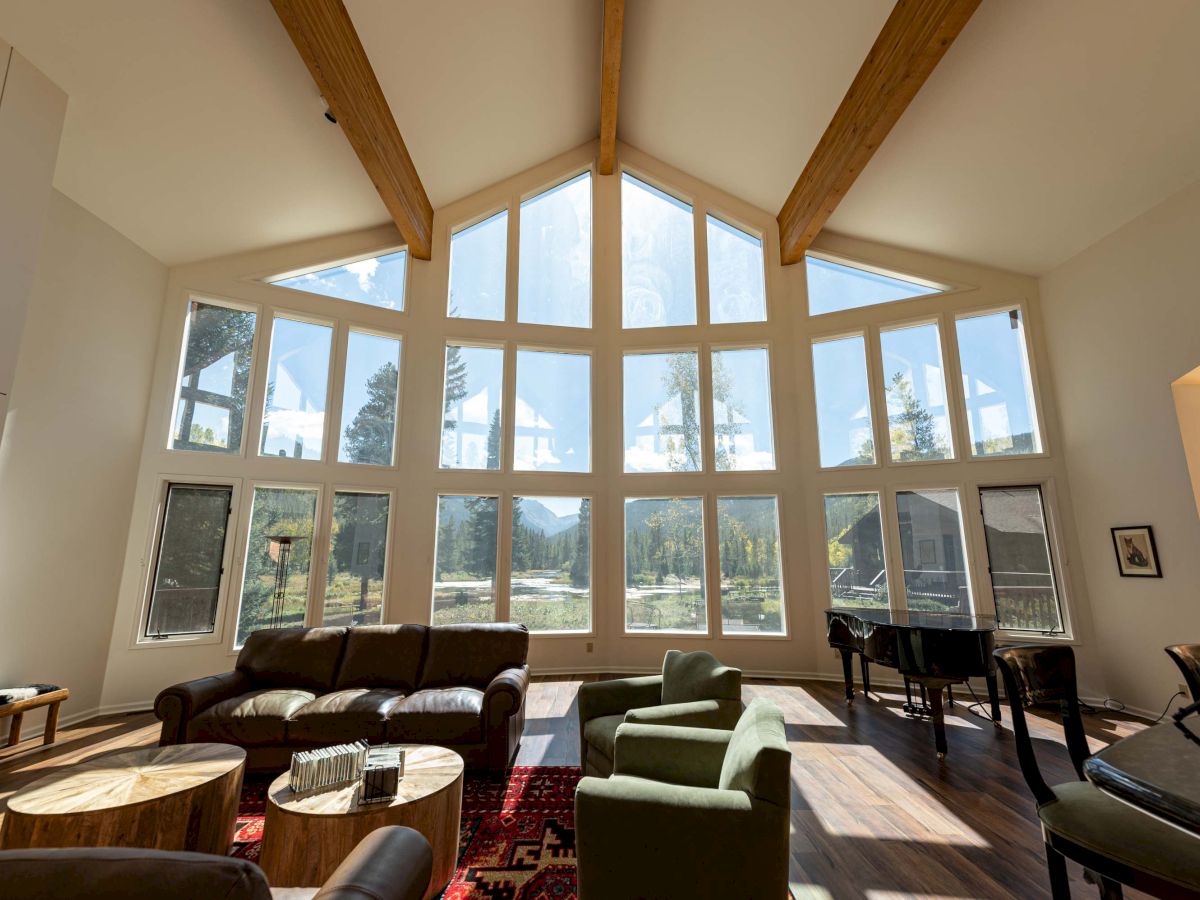 A bright, airy living room with oversized windows and a high, wooden‑beam cathedral ceiling; mid-century chairs surround circular wood tables, a piano by the windows. End sentence.