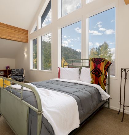 A tidy hotel bedroom with a queen bed, colorful pillows, a desk, and a large window letting in daylight.