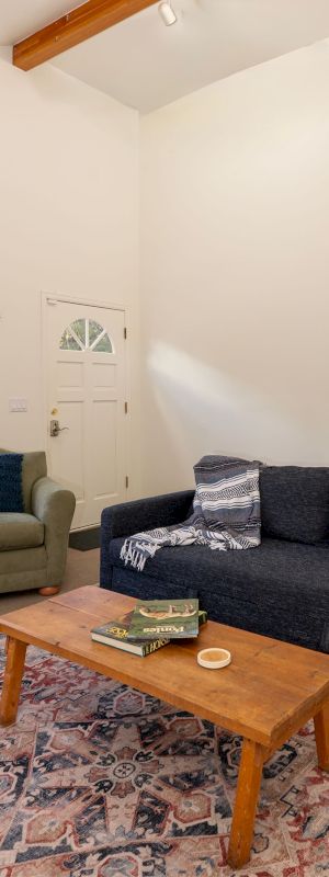 Cozy living room with two green armchairs, a dark sofa, a wooden coffee table, patterned rug, and light walls with exposed ceiling beams.