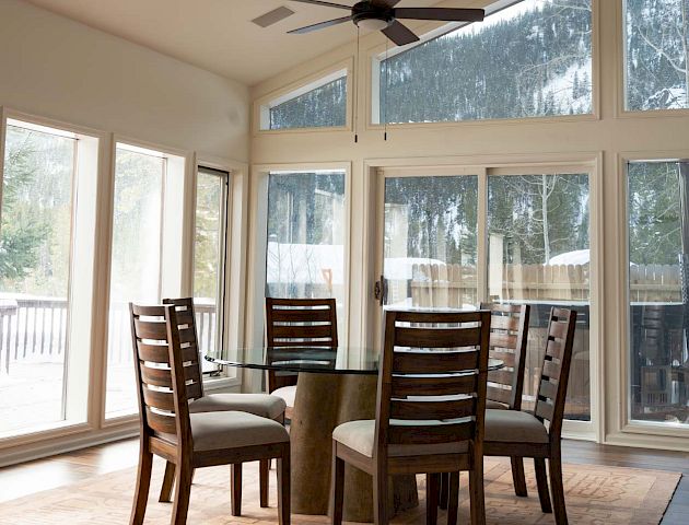 A bright sunlit dining area with a round table and four chairs, large windows, and a scenic outdoor view.