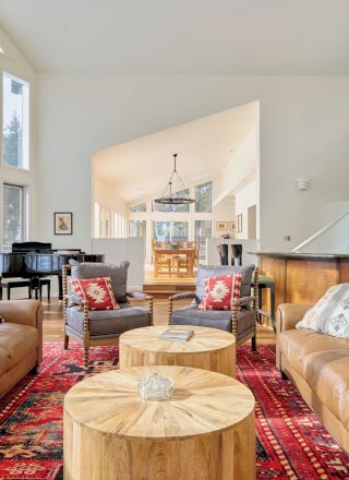 A bright, cozy living room with tan leather sofas, wooden coffee tables, a red patterned rug, and large windows letting in light.