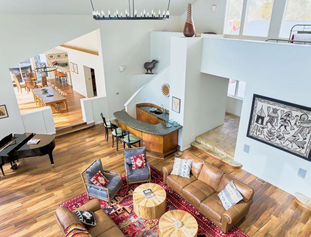 A bright, modern open-concept living area with a brown leather sofa, round wooden coffee tables, a piano, and a view into a stylish kitchen.