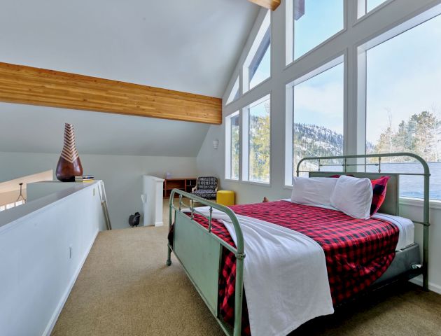 A bright loft bedroom with two metal-frame beds, plaid bedding, large windows, and a sloped ceiling overlooking a scenic view, inviting and cozy.
