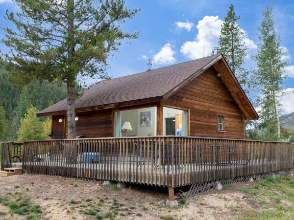 A cozy wood cabin with a large wraparound deck sits on a rocky clearing, surrounded by pines and mountains under a blue sky.