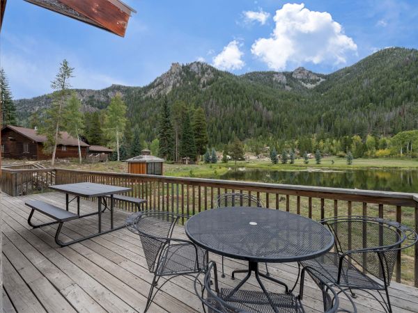 A wooden lakeside deck with round metal tables and chairs, picnic benches, a calm lake, evergreen trees, and rugged mountain peaks in the background.