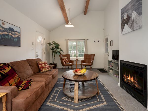 Cozy living room with a brown sectional, wooden coffee table, fireplace, area rug, wall art, potted plant, and a windowed nook with chairs.