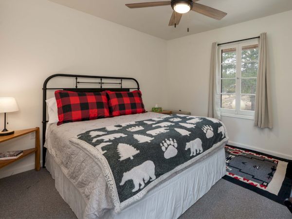 Cozy bedroom with a metal-framed bed, white bedding, a gray bear-print quilt, red plaid pillows, a small nightstand with a lamp, and a window with light curtains.