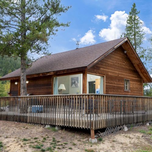 A log cabin with a wooden porch and green shutters, nestled among tall evergreen trees by a bright, sunny sky.