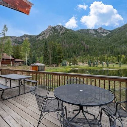 A wooden deck overlooking a lake with picnic tables and metal chairs, mountains in the background, and clear blue skies.